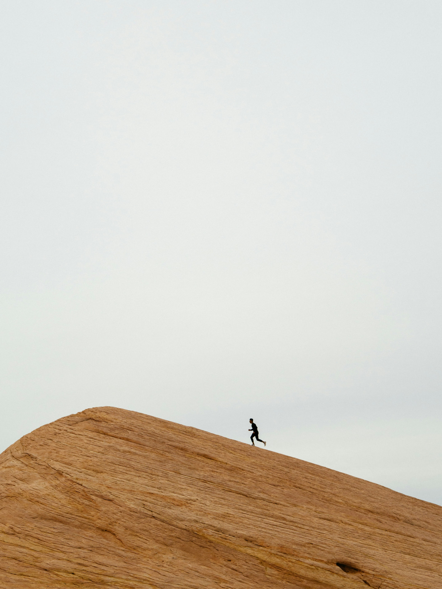 A person running up the steep face of a sand dune under a pale sky