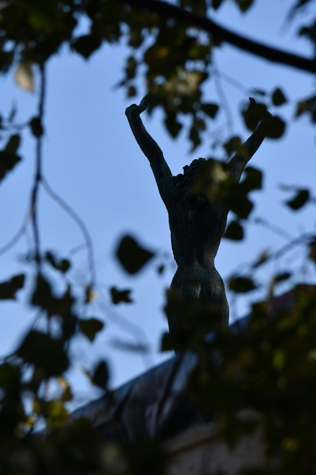 Silhouette of a statue with arms raised, seen through tree branches at dusk